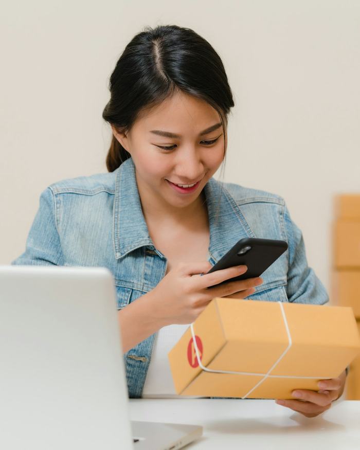 Mujer sonriendo al móvil mientras sostiene un paquete, con cajas de tienda online al fondo